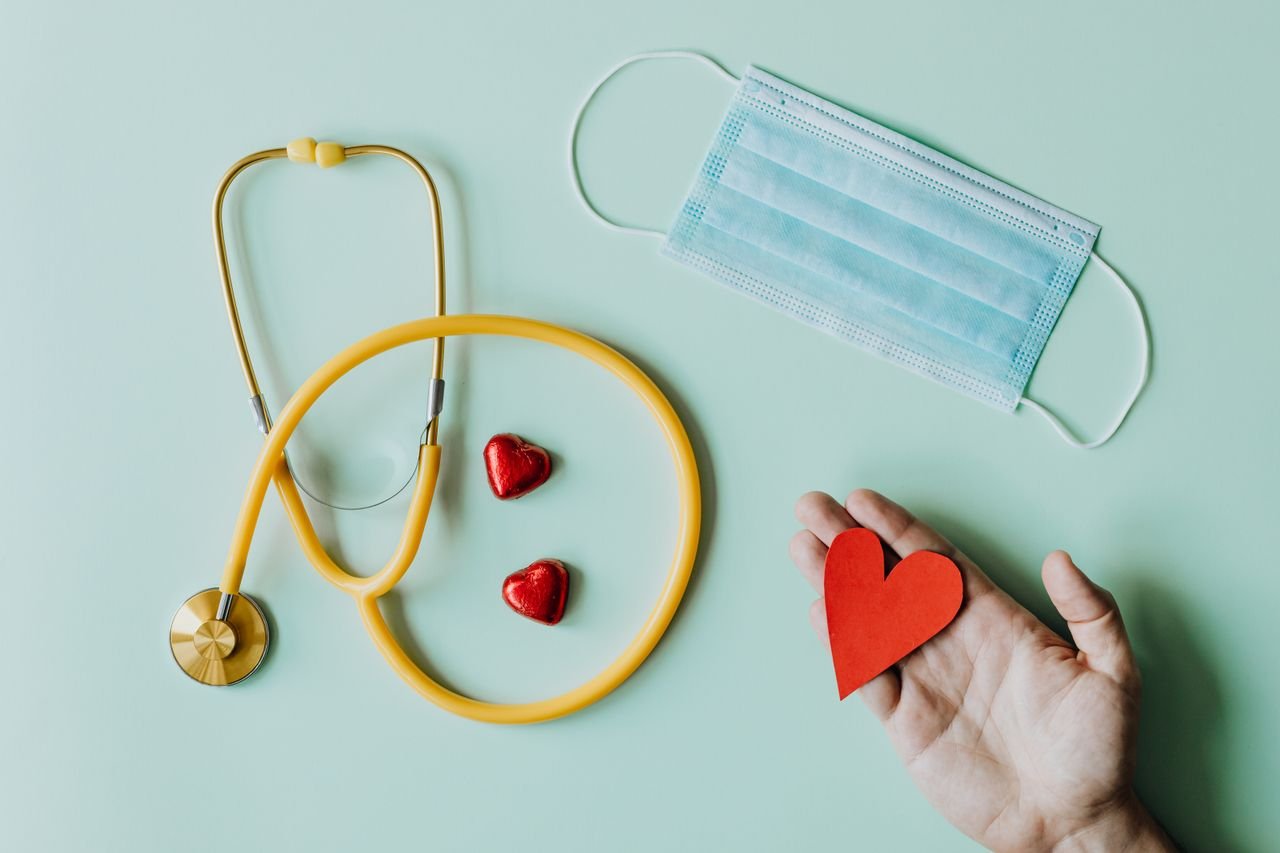 about-02 Top view of crop anonymous person hand with red paper heart on table with stethoscope and medical mask for coronavirus prevention